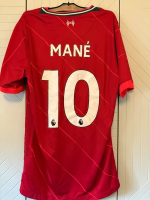 Liverpool matchtröja Mané #10 Nike - Röd Liverpool matchtröja med Mané och nummer 10 på ryggen. Tröjan har vita detaljer, klubbmärke och Nike-logga på bröstet samt sponsortryck. Tillverkad i lätt och ventilerande material, perfekt för fotboll eller träning.