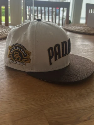 San Diego Padres keps 59FIFTY - Snygg San Diego Padres keps från New Era 59FIFTY-serien. Kepsen är vit med brunt brätte och har broderad Padres-logga samt text på sidan. Klassisk flatbrim-modell i bomullsmaterial, perfekt för dig som gillar sportig streetstyle.