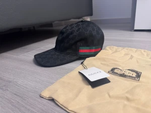 Svart canvas keps - Snygg svart keps från  med klassiskt monogram över hela kepsen. På sidan finns en ikonisk grön och röd rand. Kepsen har böjd skärm och justerbar passform. 