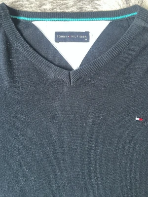 Mörkblå stickad tröja Tommy Hilfiger - Mörkblå stickad tröja från Tommy Hilfiger med V-ringning och diskret logga broderad på bröstet. Tröjan har långa ärmar och ribbade muddar vid ärmslut och nederkant. Perfekt för en stilren och klassisk look.