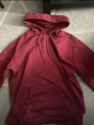 Mörkröd hoodie med magficka - Snygg mörkröd hoodie med huva och dragsnören. Klassisk modell med stor magficka framtill och ribbade muddar vid ärmslut och nederkant. Perfekt för en avslappnad och trendig stil. Materialet känns mjukt och skönt mot huden.