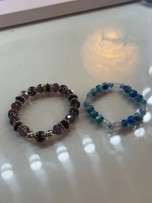 Två pärlarmband i lila och blått - Säljer två snygga armband med pärlor, ett i lila och guldtoner med svarta detaljer och ett i blå och turkosa nyanser med transparenta pärlor. Perfekta accessoarer för att ge din stil lite extra färg och personlighet.