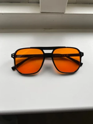 Svarta solglasögon med orange glas - Coola solglasögon med svart båge och stora, fyrkantiga orangefärgade glas. Snygg retrostil med markerad näsbrygga och breda skalmar. Perfekta för dig som vill sticka ut med en färgstark accessoar.