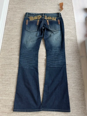 Replay blå bootcut jeans - Säljer ett par blå jeans från Replay med broderad logga i gult över bakfickorna. Jeansen har bootcut-modell, slitningar och detaljerade sömmar vid knäna. Klassisk femficksdesign och tillverkade i denim. Midjemått: 39cm rakt över, grenhöjden / midjans höjd: 18cm och innerbenets längd: 83cm