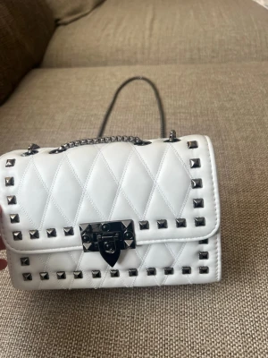 Vit axelväska med nitar och kedja - Snygg vit axelväska med quiltat mönster och silverfärgade nitar runt kanterna. Väskan har en lång kedjerem och ett coolt lås framtill. Perfekt accessoar för att lyfta din outfit med en edgy touch. Helt nytt skick oanvänd !! Pris kan diskuteras 