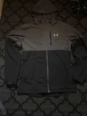 Under Armour vindjacka  - Snygg vindjacka från Under Armour i svart och grått med hel dragkedja och huva. Jackan har en bröstficka med dragkedja och logga på bröstet. Perfekt för blåsiga dagar och sportiga outfits.