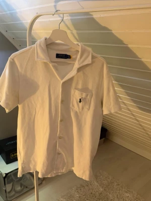 Vit kortärmad skjorta från Polo Ralph Lauren - Vit skjorta från Polo Ralph Lauren med korta ärmar, klassisk krage och knappar framtill. Bröstficka med ikonisk logga broderad i mörkblått. Tillverkad i mjuk frotté som ger en avslappnad känsla. Tröjan sitter på en kille som väger 80kg och är 190cm.