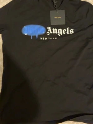 Svart Palm Angels t-shirt New York - Svart t-shirt från Palm Angels med tryck på bröstet där det står 'Palm Angels New York' i vitt och blått. T-shirten har klassisk passform och rund hals. Perfekt för dig som gillar streetwear och vill sticka ut med en snygg logga.