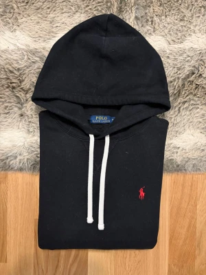 Svart Polo Ralph Lauren Fleece Hoodie - Storlek M - Hej! 👋 Säljer en riktigt snygg svart Polo Ralph Lauren hoodie i storlek M med röd pony. 🐎 Tröjan är i mycket fint skick och välvårdad. Garanterat äkta – alla tvättlappar och nacketiketter finns för verifiering. ✅ QR-koden visar just nu ”authentication check”, vilket beror på att den skannats för många gånger i tidigare annonser (Ralph Laurens säkerhetsspärr). Ett perfekt premium-basplagg som håller stilen! Fraktar snabbt! 🚛📦💬💯 