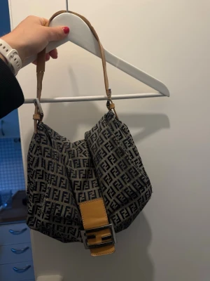 Fendi axelväska med FF-mönster - Snygg axelväska från Fendi med klassiskt FF-monogram i svart och grått tyg. Väskan har en beige läderdetalj med metallspänne framtill och matchande axelrem i läder. Perfekt för dig som gillar statement-accessoarer och ikonisk design. En spricka på bandet, den har inte gått av helt men den är trasig