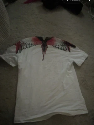 Vit t-shirt med färgglada vingar - Säljer en vit t-shirt från Marcelo Burlon med ikoniskt vingtryck i rött, svart och grått över axlar och halsringning. T-shirten har korta ärmar och en klassisk passform. Perfekt för dig som vill sticka ut med en unik design.