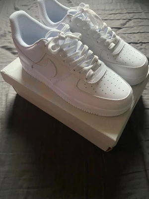 Nike Air Force 1 vita sneakers - Klassiska helvita Nike Air Force 1 sneakers med lågt skaft. Skorna har ovandel i läder, perforerad tåbox för ventilation och tydlig Nike Swoosh på sidorna. Tjock vit sula med AIR-dämpning och broderad Nike Air-logga på hälen. Perfekta för en clean och stilren look. Endast använda en gång!
