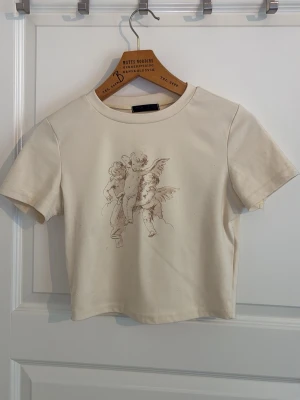 Beige croppad t-shirt med ängeltryck - Snygg beige croppad t-shirt med ett konstnärligt tryck av änglar på bröstet. T-shirten har rund halsringning och korta ärmar. Tillverkad i mjuk bomull som känns skön mot huden. Perfekt för dig som gillar unika prints och stilrena färger.