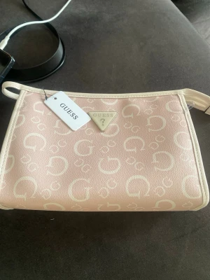Ljusrosa axelremsväska från Guess - Helt ny Snygg ljusrosa handväska från Guess med vit logotypprint över hela väskan och vita detaljer. Väskan har en klassisk rektangulär form, justerbar axelrem och Guess metallogo framtill. Materialet är konstläder som ger en lyxig känsla och trendig look.