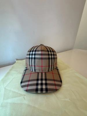 Beige keps från Burberry med rutmönster - Snygg keps från Burberry i klassiskt beige rutmönster med svarta, vita och röda linjer. En mycket fin och modern keps som är perfekt till sommaren och din outfit.
