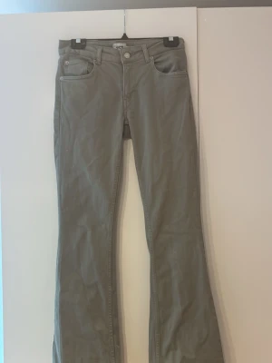gröna bootcut jeans - Säljer ett par grågröna byxor med utsvängda ben och klassisk femficksdesign. Byxorna har en enkel look utan synliga loggor och är tillverkade i ett mjukt bomullsliknande material. Perfekta för dig som gillar retro eller avslappnad stil.