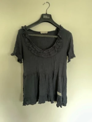 Svart blus med volang från Odd Molly - Supersöt svart blus från Odd Molly med volangdetaljer runt halsringningen och nertill. Blusen har korta ärmar och en lös passform, perfekt för en avslappnad men ändå stilsäker look. Tillverkad i mjuk bomull som känns skön mot huden.