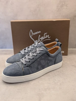 Gråa sneakers från Christian Louboutin - Stilrena gråa sneakers från Christian Louboutin i mocka med vita sulor och matchande grå skosnören. Låg modell med klassisk design och diskreta sömmar. Insidan är fodrad i beige läder för extra komfort. Perfekta för dig som gillar exklusiva och minimalistiska sneakers.