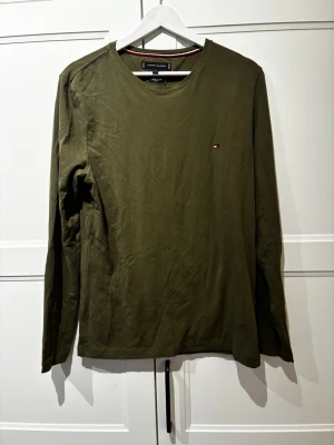 Olivgrön långärmad tröja Tommy Hilfiger - Snygg olivgrön långärmad tröja från Tommy Hilfiger i slim fit-modell. Tillverkad av ekologisk bomull och har en liten diskret logga på bröstet. Perfekt till jeans eller chinos för en clean och stilren look. Nypris (600)