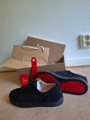 Svarta sneakers från Christian Louboutin - Snygga svarta sneakers från Christian Louboutin i mocka med klassisk röd sula. Skorna har lågt skaft, svarta skosnören och diskret branding. Perfekta för dig som vill ha en stilren och lyxig look med en ikonisk touch.