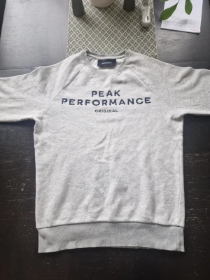 Grå sweatshirt från Peak Performance - Snygg grå sweatshirt från Peak Performance med mörkblå text på bröstet. Klassisk rund halsringning och ribbade muddar vid ärmslut och nederkant. Perfekt för en avslappnad och sportig stil. Dock har den ett litet hål på baksidan av vänster ärm men är inte synligt.