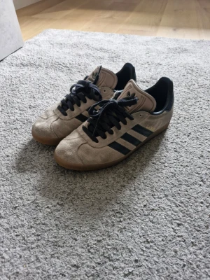 Adidas Gazelle  - Adidas Gazelle sneakers i beige mocka med svarta detaljer och klassiska tre ränder på sidorna. Skorna har svart snörning, låg profil och gummisula. Perfekta för en stilren och sportig look. Hör av er vid frågor