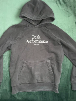 Svart hoodie Peak Performance - Snygg svart hoodie från Peak Performance med stor logga och text 'Est. 1986' på bröstet. Klassisk känguruficka framtill och huva med dragsko. Perfekt för en avslappnad och sportig stil. Mjuk bomullskänsla och långa ärmar.