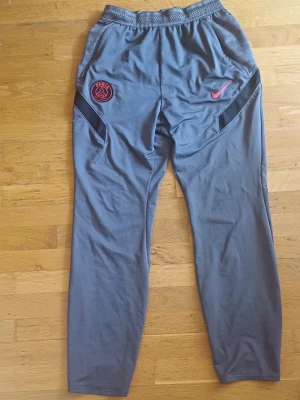 Gråa PSG x Nike träningsbyxor - Säljer ett par gråa träningsbyxor från Nike i samarbete med PSG. Byxorna har elastisk midja, svarta detaljer vid fickorna och PSG-logga samt Nike Swoosh i rött framtill. Perfekta för fotboll eller gymmet. Storlek 158 - 170. Byxorna är äkta. Byxorna har ett litet hål men annars är dom i perfekt skick. 