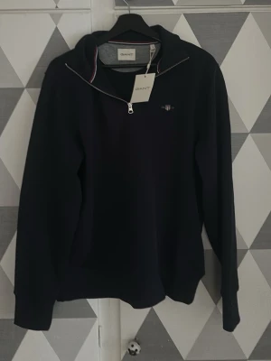 Svart halvzip tröja från GANT - Stilren svart långärmad tröja från GANT med halvzip och liten broderad logga på bröstet. Tröjan har en klassisk krage med dragkedja och diskreta ränder på insidan av kragen. Perfekt för en avslappnad men snygg look. Den är ny och aldrig använd med lappen kvar.