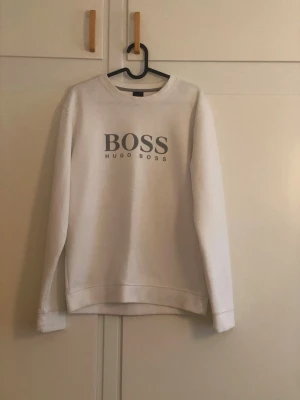 Vit sweatshirt från Hugo Boss - Vit sweatshirt med rund hals och tryckt BOSS-logo på bröstet. Tröjan har en klassisk passform med långa ärmar och ribbade muddar vid ärmslut och nederkant. Perfekt för en clean och stilren look.
