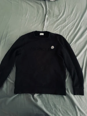 Svart sweatshirt från Moncler - Svart långärmad sweatshirt från Moncler med diskret logotyp på bröstet och ett broderat Moncler-märke på vänster sida. Klassisk rund halsringning och mjukt material som ger en clean och stilren look.