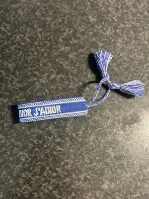 Blå armband Dior - Snyggt blått armband från Dior med texten 'DIOR J'ADIOR' och 'CHRISTIAN'. Armbandet är vävt i tyg och har tofsar i ändarna för en trendig look. Perfekt accessoar för att lyfta din outfit med en lyxig touch.