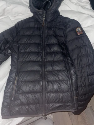 Svart dunjacka från Parajumpers - Snygg svart dunjacka från Parajumpers med huva och dragkedja framtill. Jackan har två sidofickor med dragkedja och ett emblem på ena ärmen. Perfekt för kalla vinterdagar och riktigt skön att bära. Helt ok skick men håller en varm och fin.