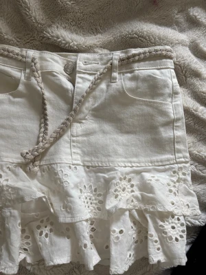 Vit kjol med volang och broderi - Superfin vit kortkjol med volangkant och broderade blommor. Kjolen har fickor fram, flätat bälte och är gjord i bomull. Perfekt för en fräsch och somrig look! Kjolen är stor i storleken 