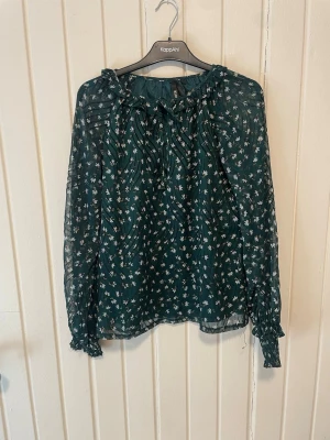 Grön blommig blus från Elli White - Mörkgrön transparent blus från Elli White med små vita och rosa blommor. Blusen har långa ärmar med resår vid ärmslut och volangdetaljer vid halsen. Tillverkad i ett lätt och luftigt material som ger en flowy känsla.