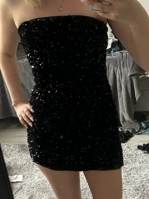 Svart glitterklänning axelbandslös - Säljer en svart, axelbandslös kortklänning täckt med glittriga paljetter. Klänningen är ganska kort,(jag är 170). Använd en gång.