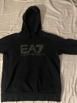 Armani hoodie - Snygg svart hoodie från EA7 Emporio Armani med stor logga på bröstet. Klassisk känguruficka framtill och justerbar huva med snören. Tillverkad i mjukt bomullsmaterial som är skönt mot huden. Perfekt för en avslappnad och trendig look.