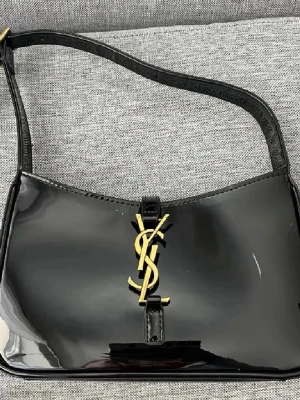 Svart YSL axelväska i lackläder - Snygg svart axelväska från Yves Saint Laurent i glansigt lackläder med guldigt YSL-logo framtill. Väskan har en stilren, rektangulär form och ett smalt axelband. Perfekt för dig som vill ha en trendig och lyxig accessoar med ikonisk design.