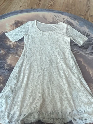 Vit spetsklänning med kort ärm - Söt vit klänning i spets med rund halsringning och korta ärmar. Klänningen har ett fint blommigt spetsmönster och är lätt utsvängd i modellen. Perfekt för sommarens alla tillfällen.