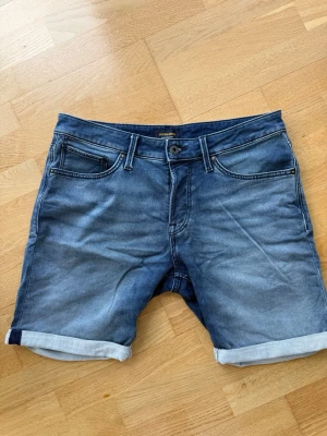 Blå denimshorts från Jack & Jones - Snygga blå denimshorts från Jack & Jones med klassisk femficksdesign och uppvikta benslut. Shortsen har normal passform och stängs med knapp och dragkedja. Perfekta för varma dagar och enkel att matcha med t-shirt eller skjorta.