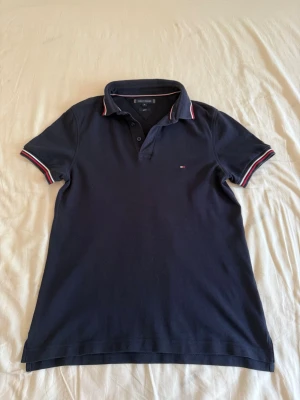 Tommy Hilfiger Piké Slim Fit - Klassisk mörkblå pikétröja från Tommy Hilfiger med korta ärmar och krage. Tröjan har diskret logga på bröstet och rödvita detaljer på krage och ärmslut. Perfekt för en stilren och sportig look.