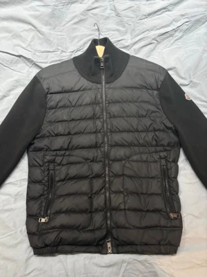 Svart cardiganjacka från Moncler - Snygg svart cardiganjacka från Moncler med quiltad framsida och ärmar i stickad ull. Jackan har hög krage, två dragkedjefickor och Moncler-logga på ärmen. Perfekt för lager-på-lager och streetstyle.