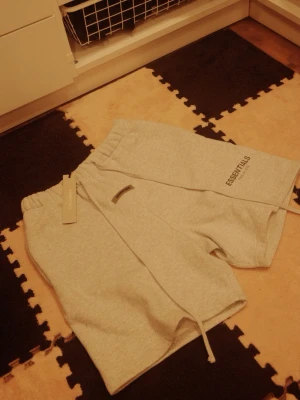 Grå Essentials shorts med snörning - Snygga grå shorts från Essentials med elastisk midja och snörning framtill. Mjukt material och logga tryckt på ena benet. Perfekta för en avslappnad och trendig stil.