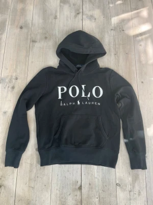 Svart hoodie från Polo Ralph Lauren - Svart hoodie från Polo Ralph Lauren med stor vit logga på bröstet. Klassisk känguruficka framtill, justerbar dragsko i huvan och ribbade muddar vid ärmar och midja. Tillverkad i mjukt bomullsmaterial för en skön och avslappnad look. Passar dig som är va 165-170 cm