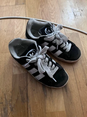 Adidas Campus svarta sneakers - Svarta Adidas Campus sneakers med klassiska vita ränder och vit sula. Skorna har gråvita skosnören och är låga i modellen. Ovandelen är i mocka och insidan är ljusgrå. Perfekta för en avslappnad streetstyle-look.