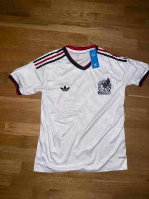 Vit Mexiko Adidas tränings tröja vm2026 - Säljer en vit retro fotbollströja från Adidas med Mexikos landslagsmärke på bröstet. Tröjan har svarta och röda detaljer vid krage och ärmslut samt klassiska Adidas-ränder på axlarna. Tillverkad i lätt och ventilerande material, perfekt för träning eller match.