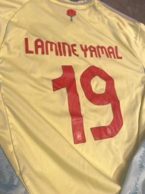 Spanien em tröja 2024 Yamal 19 - Snygg gul fotbollströja från Spanien med Lamine Yamal och nummer 19 på ryggen. Tröjan har röda detaljer, tryck på ryggen och UEFA Foundation-märke på ärmen. Tillverkad i lätt och ventilerande material, perfekt för match eller träning. Äkta adidas 