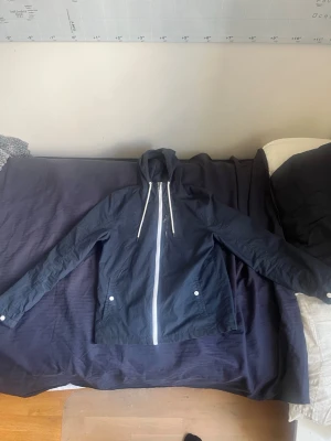 Mörkblå vindjacka med huva - Snygg mörkblå vindjacka med vit dragkedja och vita snören i huvan. Jackan har två fickor med knappar och är tillverkad i ett lätt material som passar perfekt för blåsiga dagar. Enkel och stilren design som funkar till det mesta.Obs super litet hål i armhålan 