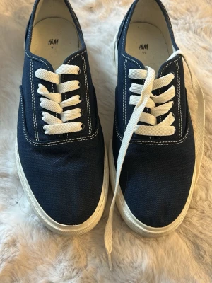 Mörkblå sneakers från H&M - Snygga mörkblå sneakers från H&M med vit sula och vita skosnören. Skorna är i canvasmaterial och har klassisk låg modell med kontrasterande sömmar. Perfekta för en avslappnad och stilren look. Använda 2 gånger