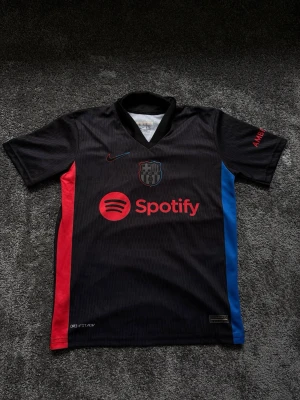Svart FC Barcelona matchtröja Nike - Snygg svart FC Barcelona matchtröja från Nike med röda och blå detaljer på sidorna. Tröjan har klubbmärke och Nike-logga på bröstet samt Spotify som sponsor framtill. Tillverkad i lätt och ventilerande material, perfekt för fotbollsträning.